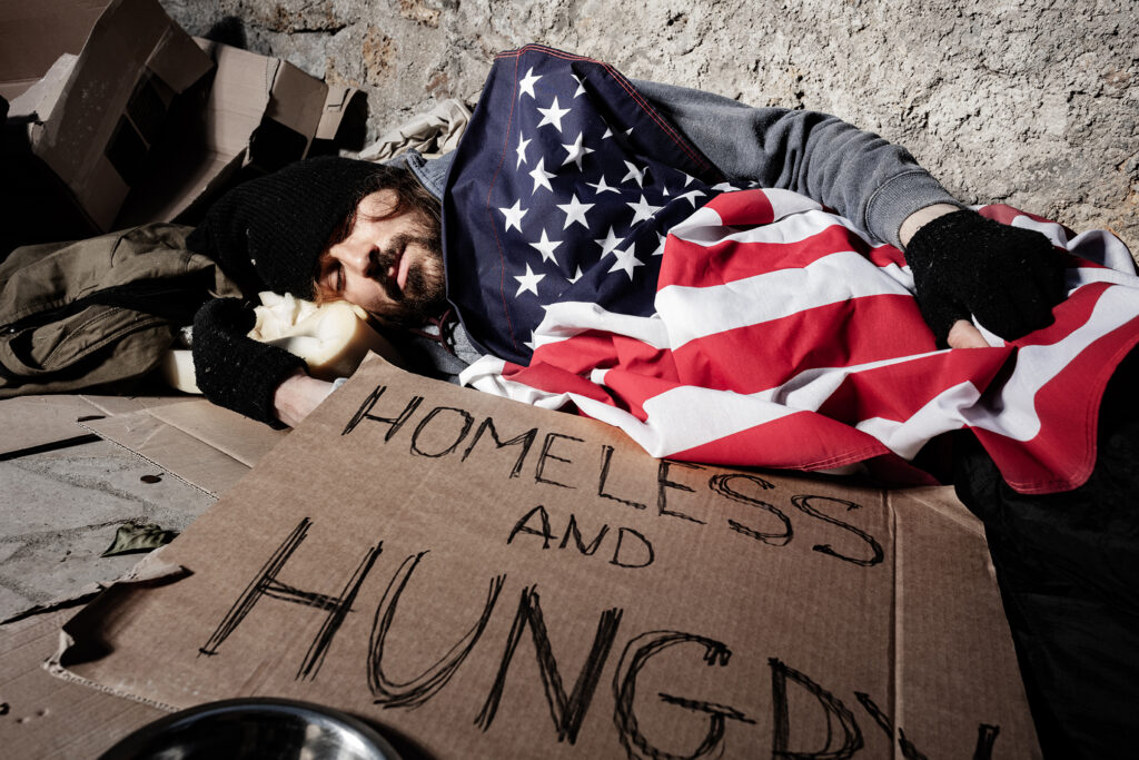 Homeless man draped in an American flag, asleep on the street with sign, "Homeless and Hungry"