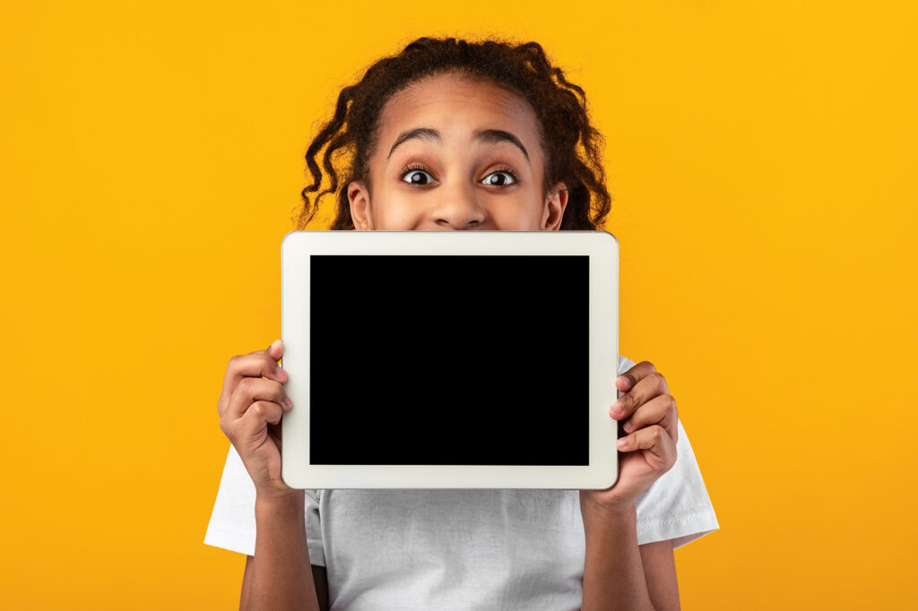 Great Application. Excited Black Girl Holding And Showing Digital Tablet With Empty Screen For Mockup, Covering Face And Mouth, Recommending New App Posing Standing Over Yellow Studio Background